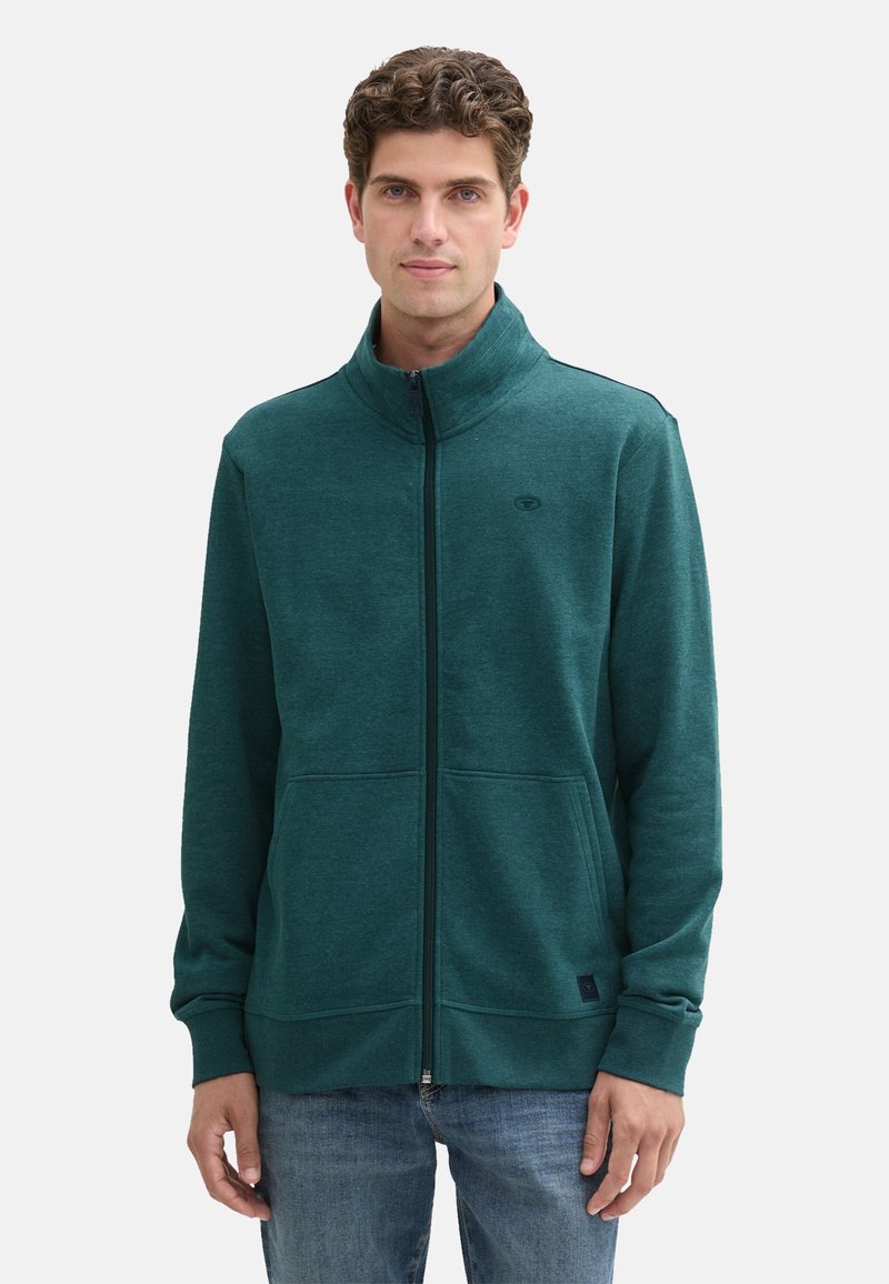 Giacca verde con zip realizzata in tessuto morbido, dotata di un alto colletto, due tasche frontali e un logo sul lato sinistro del petto.