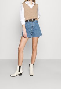 Suéter beige sin mangas de punto, camisa blanca con cuello debajo, pantalones cortos de mezclilla azul, cinturón negro con tachuelas, botines blancos con detalles negros.