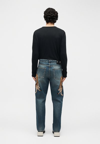 Άτομο με σγουρά μαλλιά που φοράει μαύρο μακρυμάνικο πουκάμισο και μπλε τζιν με κέντημα τίγρης στις πίσω τσέπες, στέκεται με την πλάτη γυρισμένη.