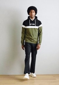 Grønn og svart hettegenser med hvitt stripe, med en logo. Kombinert med svarte bukser og hvite joggesko; modellen står foran en nøytral bakgrunn.