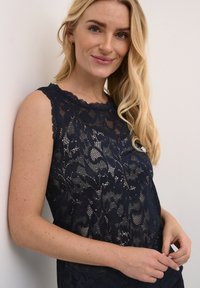 Jūras zils mežģīņu topiņš ar ziedu rakstiem, viļņotām kakla malām un bez piedurknēm. Caurspīdīga tekstūra ar pieguļošu siluetu.