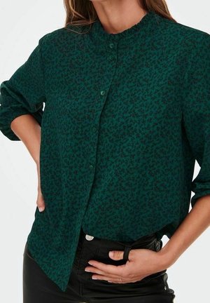 Femme portant une chemise boutonnée à manches longues vert foncé avec un motif floral subtil, associée à un pantalon noir.