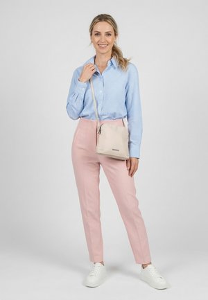 Frau lächelt, steht und trägt ein blau kariertes Hemd, pinke Hosen, weiße Turnschuhe und eine beige Umhängetasche.