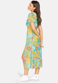 Tropenmuster, kurzärmliges Kleid in Blau, Gelb und Pink, mit fließendem Schnitt und seitlichem Schlitz. Kombiniert mit weißen Sneakers.