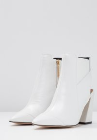 Botas de tobillo de cuero blanco con puntas afiladas, cremallera lateral y tacón grueso. Presentan un diseño elegante y una textura suave. Detalle de herrajes dorados.