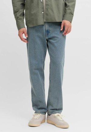 Homme portant un jean bleu clair ample, des baskets beiges et une chemise à carreaux verte boutonnée, debout devant un fond uni.