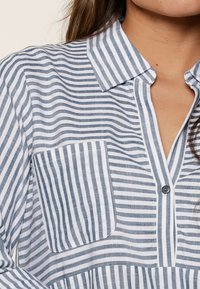 Camisa de botones a rayas en azul y blanco, con cuello, bolsillo frontal y textura suave, con rayas verticales y horizontales.