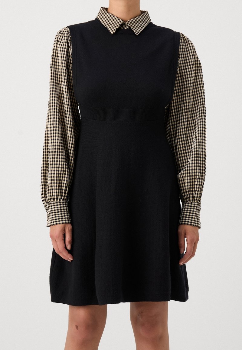 Femme portant une robe noire sans manches par-dessus une chemise à manches longues à carreaux beige et noirs avec col, debout devant un fond blanc.