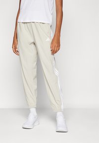 Helle beige Sporthosen mit weißen Seitenstreifen, elastischen Bündchen und einer lockeren Passform, kombiniert mit weißen Sportschuhen.