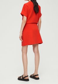 Conjunto de dos piezas rojo que incluye una blusa de manga corta y una falda acampanada. El material tiene una textura suave. Combinado con sandalias negras de tiras.