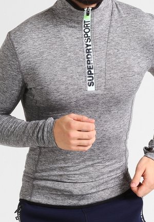 Mand iført tætsiddende grå langærmet lynlåstrøje med teksten "Superdry Sport" på lynlåsen, i en løbestilling.