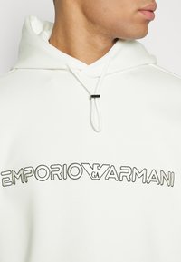 Vit huvtröja med dragsnodd, med svart broderad "EMPORIO ARMANI"-logotyp på framsidan i ett djärvt, modernt teckensnitt.