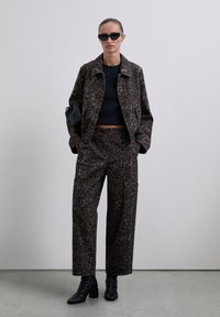 Woman wearing dark brown leopard print jacket and pants, black cropped top, black boots, and sunglasses, standing against a plain wall.