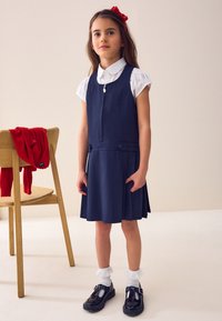 Navyblaues Kleid mit Reißverschluss, seitlichen Knöpfen und plissiertem Rock; kombiniert mit einem weißen Hemd mit Kragen. Schwarze Lackschuhe und frilly weiße Socken.