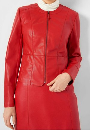 Kobieta ubrana w dopasowaną czerwoną skórzaną kurtkę na zamek, do tego pasującą czerwoną skórzaną spódnicę i biały golf.