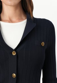 Námořnický žebrovaný cardigan vyrobený z texturovaného materiálu, s klopovým límcem, zlatými knoflíkovými detaily a malou náprsní kapsou.
