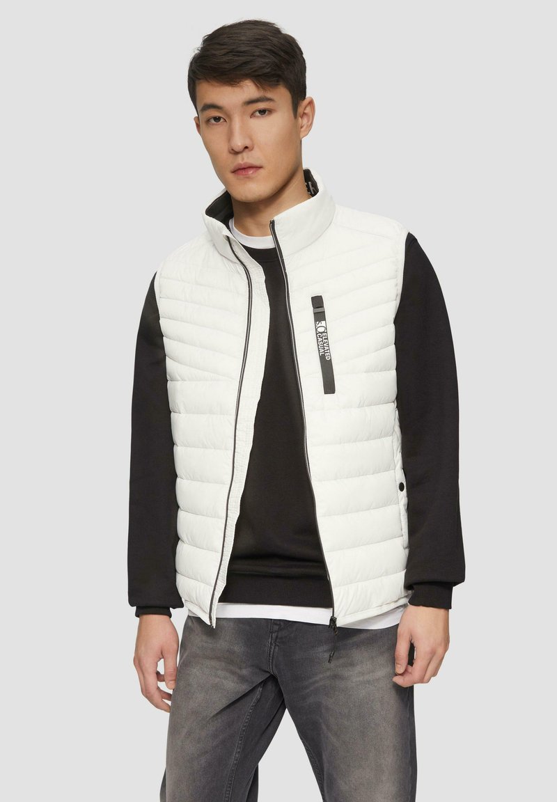 Weißer gepolsterter Weste mit hohem Kragen und schwarzem Reißverschluss, getragen über einem schwarzen Pullover. Graue Jeans und ein neutraler Hintergrund runden das Outfit ab.