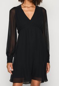 Schwarzes Kleid aus leichtem Stoff mit V-Ausschnitt, langen transparenten Ärmeln und lockerem Sitz mit gerafften Details an der Taille.