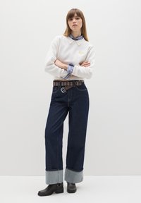 Weißes Sweatshirt mit Kragen und gesticktem Design, blau-weiß gestreiftes Hemd darunter, dunkle Jeanshose, brauer Gürtel mit Ösen, schwarze Stiefel.