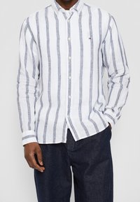 Camicia in lino bianco con righe verticali blu navy, colletto con bottoni e maniche lunghe; indossata con pantaloni in denim scuro, con vestibilità comoda.