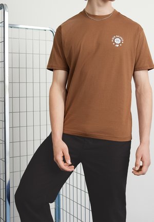 Persona que lleva una camiseta marrón de New Balance y pantalones negros, de pie junto a una división de rejilla metálica contra una pared lisa y clara.