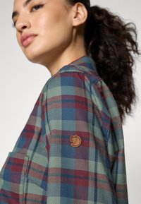 Plaid marškiniai žaliais, mėlynais ir bordo atspalviais; turi kišenę ir smėlio spalvos logotipą ant rankovės. Minkšta tekstūruota medžiaga.