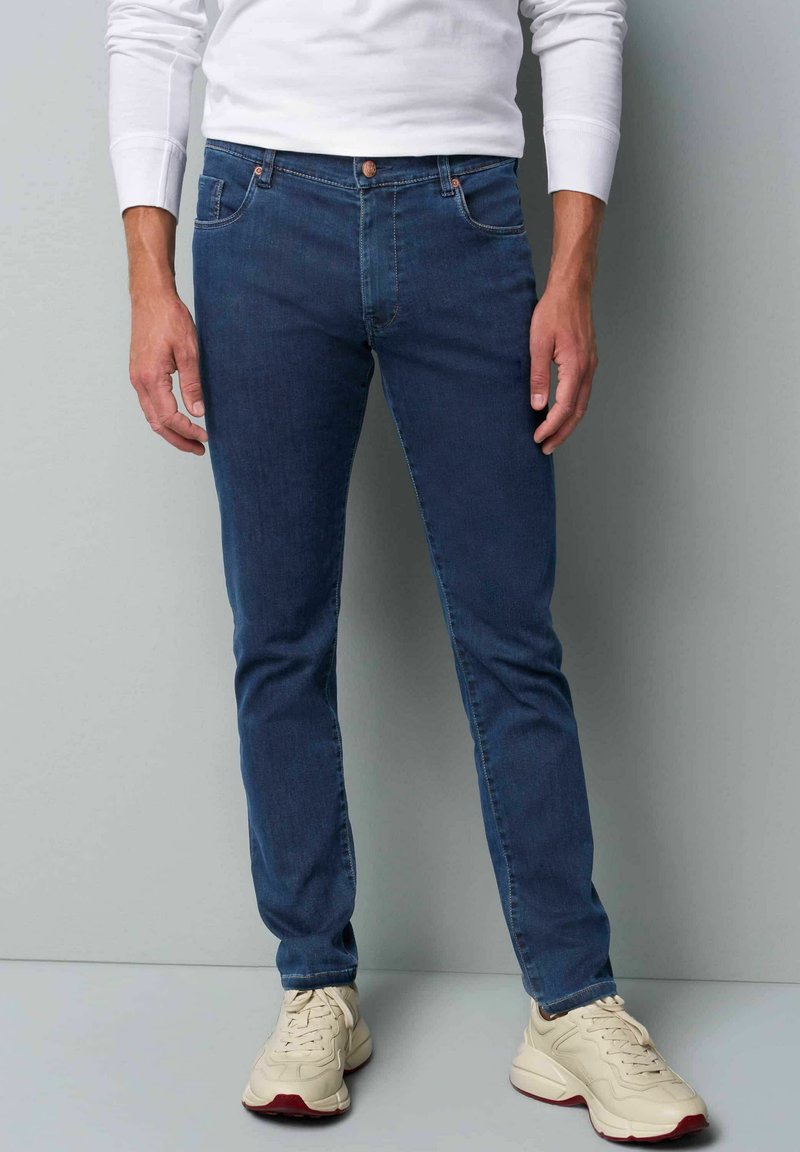 Jean slim bleu foncé avec un design classique à cinq poches, fermeture à bouton en métal, couture discrète, porté avec des baskets de couleur claire.
