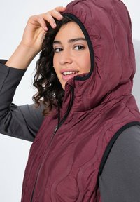 Burgunderfarbene gestepptes Weste mit Kapuze und schwarzem Rand, ausgestattet mit einem Frontreißverschluss und einem wellenförmigen Muster; getragen über einem grauen Langarmoberteil.