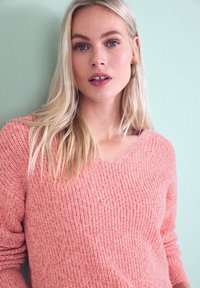 Femme blonde aux yeux bleus portant un pull en maille rose, debout devant un mur vert clair, regardant droit devant avec une expression neutre.