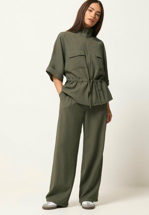 Femme debout portant un pantalon large vert olive et une veste zippée à manches courtes avec une taille à cordon de serrage et des poches plaquées, portant des baskets blanches.