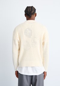 Cárdigan de punto crema con un patrón texturizado, dobladillo y puños de canalé, y un corte holgado. Llevado sobre una camisa blanca y pantalones grises.