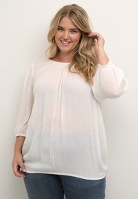 Hvid, let bluse med teksturerede polka dots, rund halsudskæring og 3/4 puffede ærmer. Båret over denimjeans.