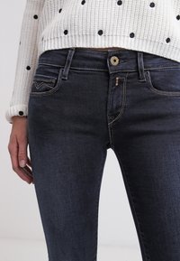 Los jeans de mezclilla oscura presentan un diseño ajustado, cinco bolsillos y costuras en contraste. Combinados con un suéter blanco texturizado con lunares negros.