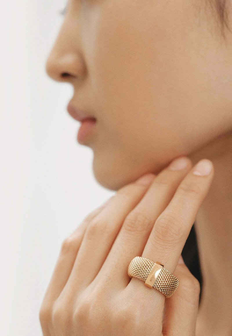 Anillo de malla de oro con una banda lisa y pulida. El diseño presenta un patrón texturizado y un acento central liso, destacando su artesanía.