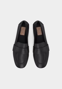 Ζεύγος μαύρων δερμάτινων μοκασίνων με φαρδιά λουριά και ετικέτα της μάρκας AGL στο εσωτερικό, τοποθετημένα δίπλα-δίπλα σε λευκό φόντο.