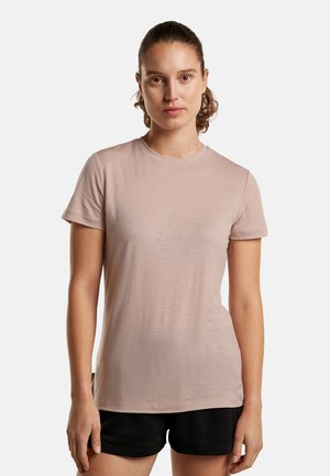 Woman with tied-back hair wearing a light pink short-sleeve t-shirt and black shorts, standing against a plain white background.