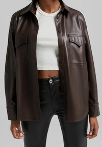 Brun faux läder skjorta med knappar och krage, två bröstfickor och en avslappnad passform, ihop med en vit ribbad croppad topp.