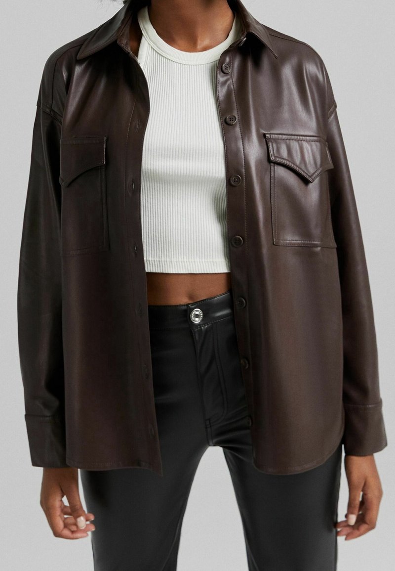 Brun faux läder skjorta med knappar och krage, två bröstfickor och en avslappnad passform, ihop med en vit ribbad croppad topp.