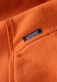 Tela de algodón naranja con una textura suave, que presenta una etiqueta azul marino cosida con la palabra "SUPERDRY" en costura blanca.