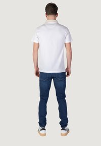 Camisa polo blanca con cuello crema, combinada con jeans azules y zapatillas blancas con detalles en negro. Costuras visibles en la ropa.