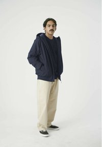 Marineblaue leichte Jacke mit Kapuze, Reißverschluss vorne und elastischen Bündchen, kombiniert mit beigen weit geschnittenen Hosen und schwarzen Sneakers.