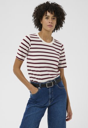 Femme aux cheveux bouclés portant un t-shirt à manches courtes blanc à rayures bordeaux, rentré dans un jean taille haute bleu avec une ceinture noire tressée.