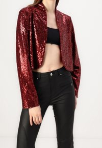 Red sequin cropped jacket with long sleeves, rounded collar, and smooth black tank top, paired with fitted black pants featuring side pockets.