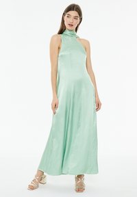 Abito maxi in satin verde menta con collo alto, senza maniche e spacchi laterali. Abbinato a sandali con tacco dorati.