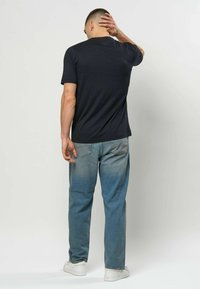 Mann i svart t-skjorte og blå jeans, stående med ryggen til kameraet, høyre hånd på nakken, hvite joggesko synlige på ensfarget bakgrunn.