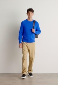 Maglione blu lavorato a maglia, chinos beige e sneaker nere. Il modello tiene una borsa scura, su sfondo di pareti texture, in un contesto neutro.