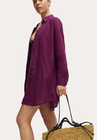 Lila, langärmliges Button-Down-Hemd aus leichtem Stoff, mit einem leicht geschwungenen Saum, über einem dunklen Oberteil getragen, kombiniert mit einer geflochtenen Tasche.