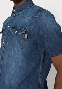 Koszula dżinsowa z krótkim rękawem w ciemnym odcieniu niebieskiego, z podwójnymi kieszeniami na klatce piersiowej z klapami, kołnierzykiem zapinanym na guziki oraz kontrastującymi detalami w stębnowaniu.