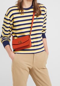 Koszula w paski z długim rękawem w kolorach granatowym, żółtym i białym, zestawiona z pomarańczą torebką na ramię z klapką i tłoczonym logo. Beżowe spodnie.
