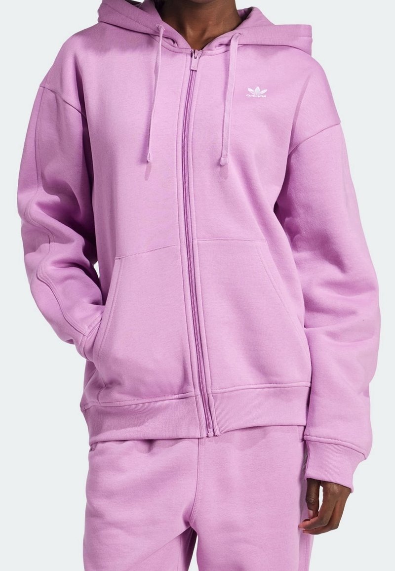 Osoba oblečená do světle fialové mikiny na zip s klokankovými kapsami a odpovídajícími tepláky, logo Adidas viditelné na hrudi.